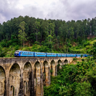 A blue train crossing Nine Arches Bridge in Ella, Sri Lanka, against the backdrop of mountains covered in vibrant green foliage.